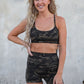 Woman wearing a camouflage sports bra and shorts set standing against a concrete wall.